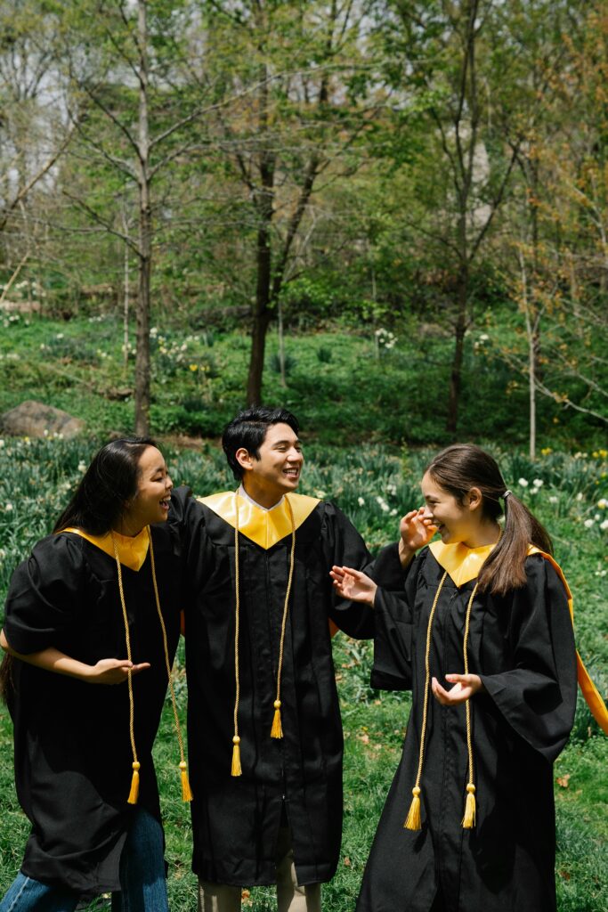 Three smiling graduates in black gowns celebrate outdoors surrounded by lush greenery.